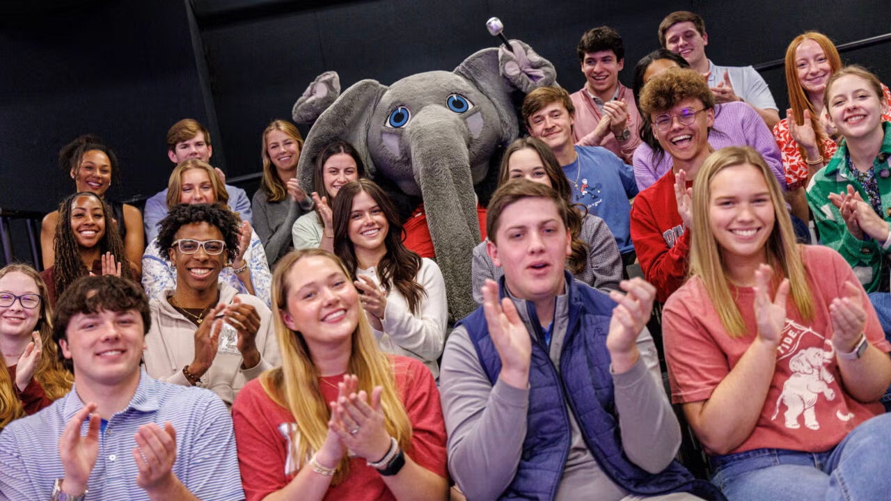 A group of college students and a costumed elephant cheer in a studio