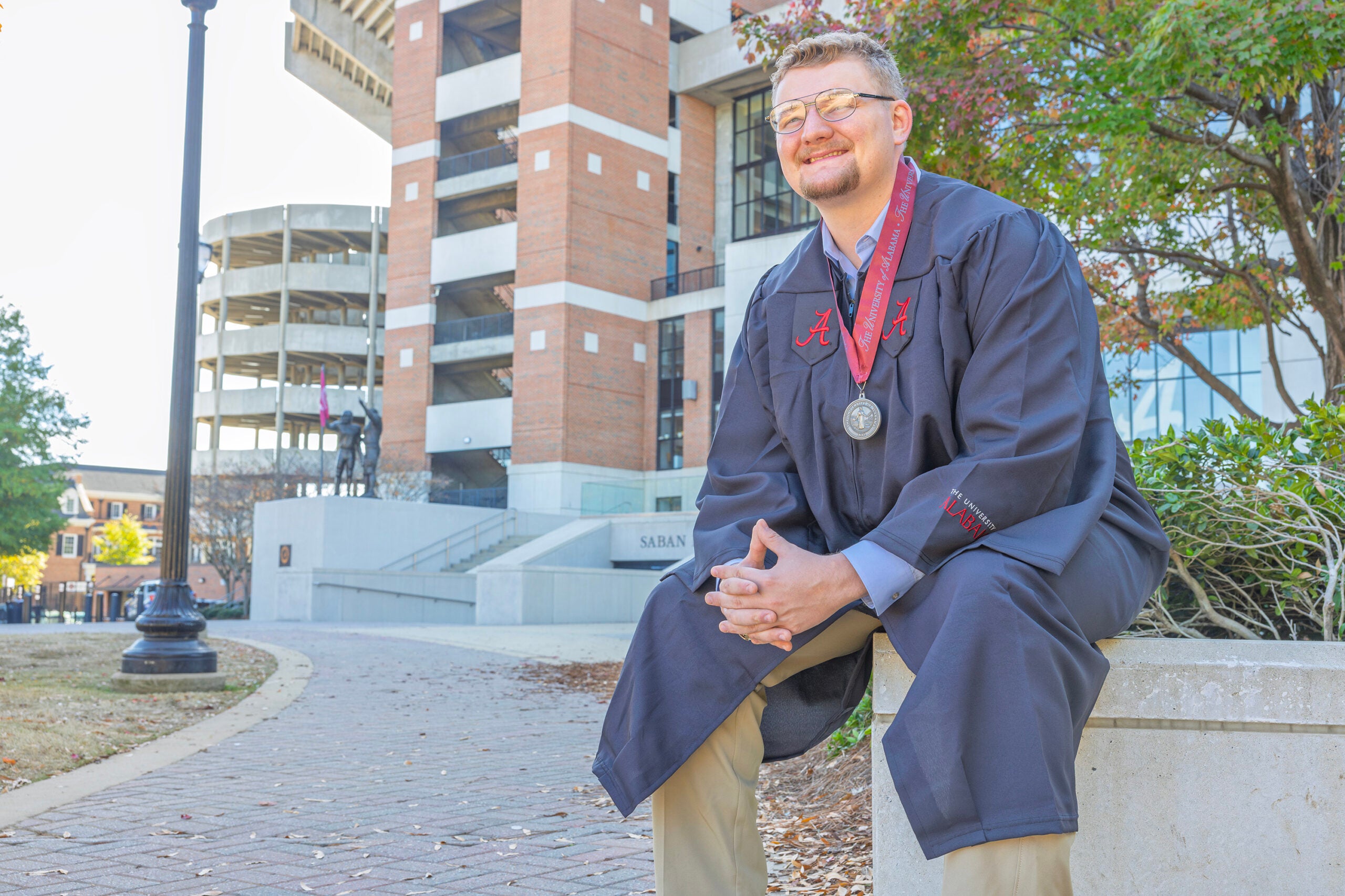 a male college student wearing a commencement gown sits on a bench beside a sports stadium