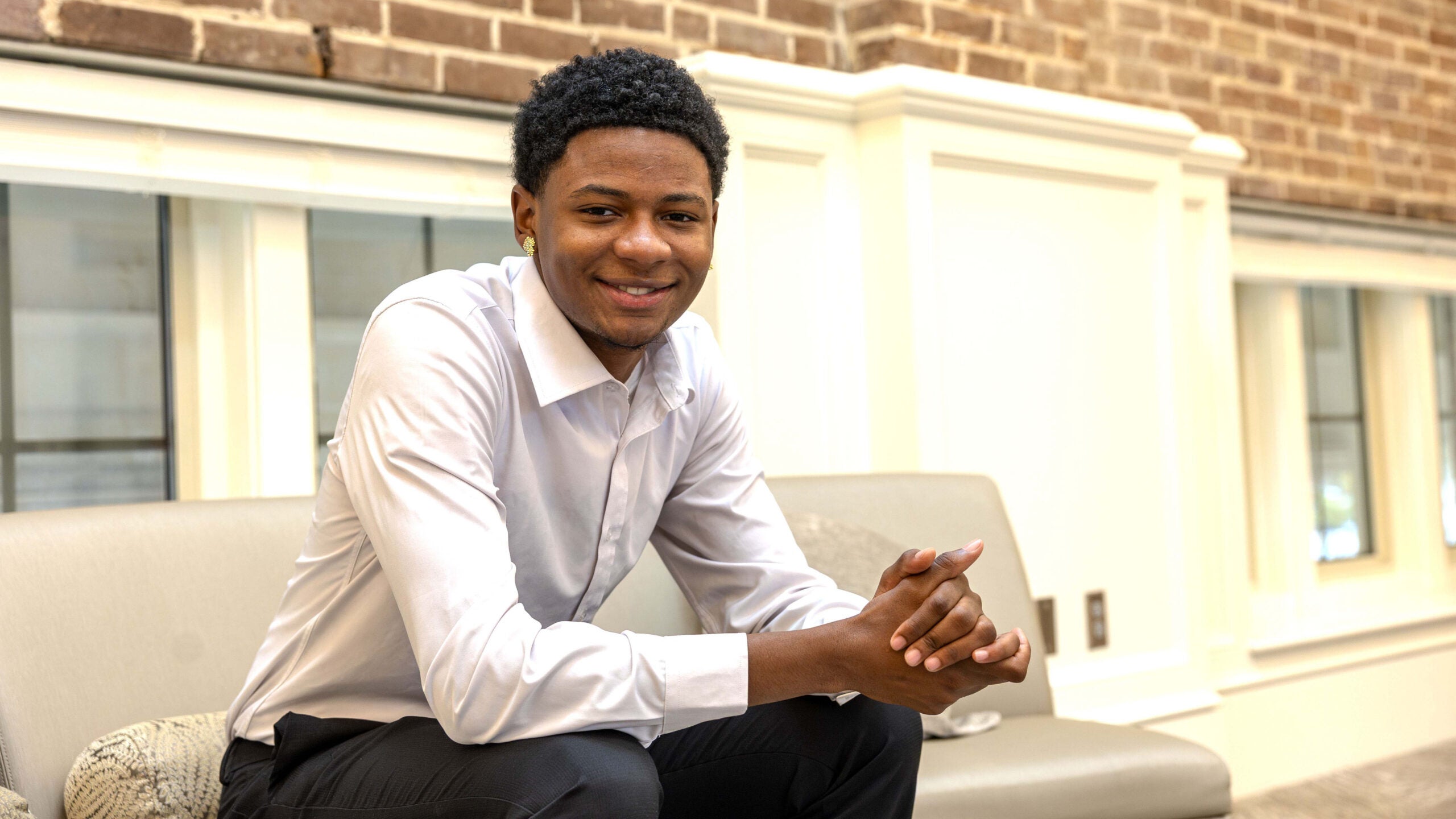 A male college student sits on a couch near a window and poses for a photo.