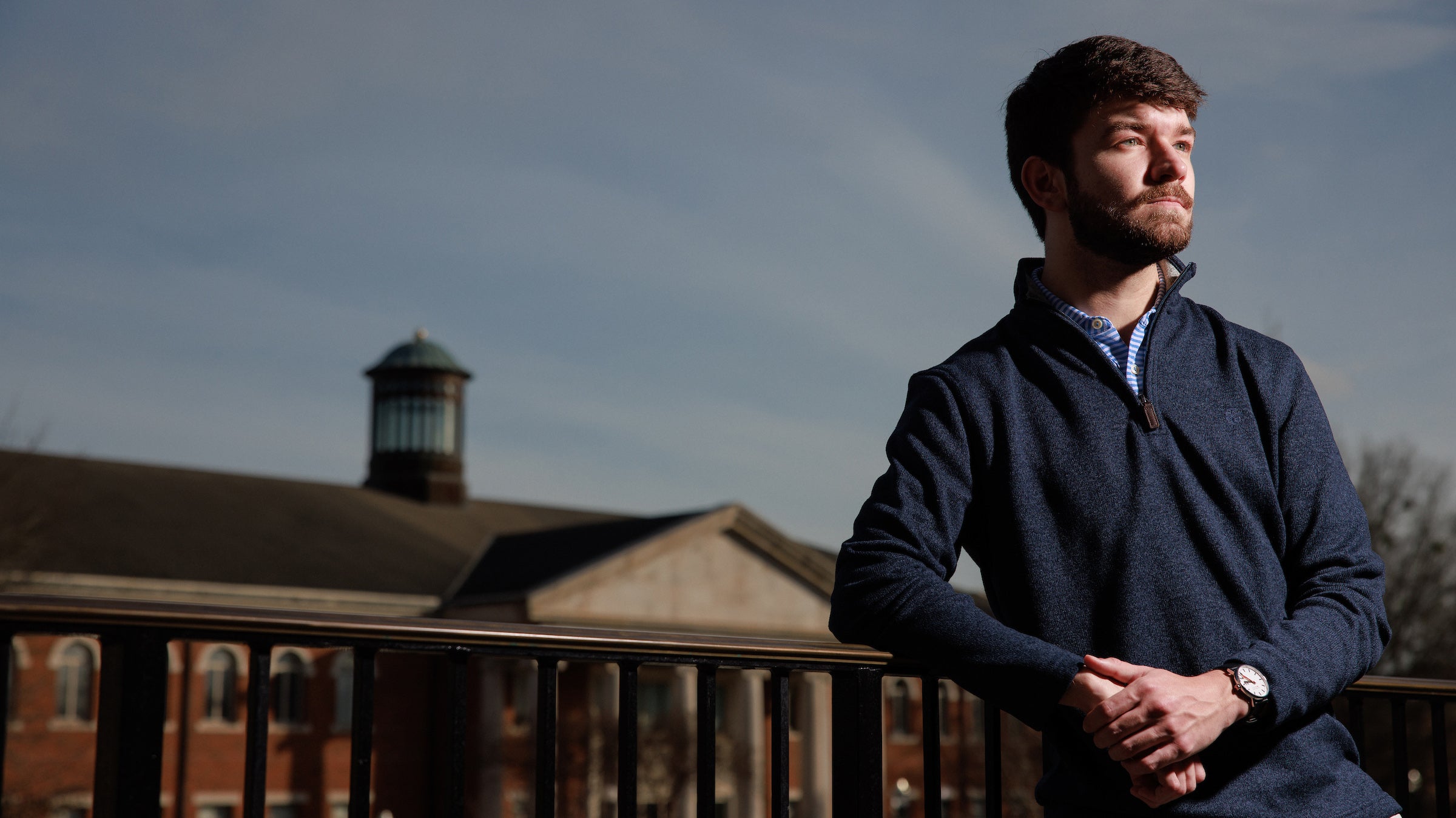 A male college student leans on a balcony railing and poses for a photo