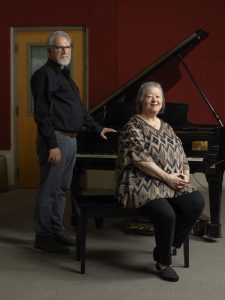 A white man and white woman pose for a photo next to a piano