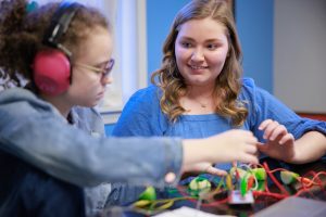 A white female graduate student explains a robotics task to a young girl