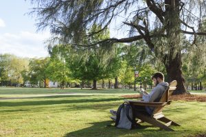 A white male college studen reads a book while sitting in a chair outdoors